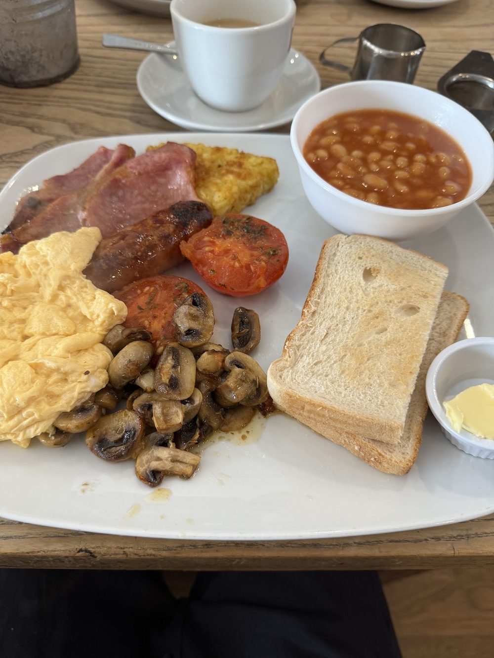 Cooked breakfast of bacon, sausage, scrambled egg, mushrooms, grilled tomato, hash brown, toast and a bowl of baked beans, with a cup of tea and a milk jug on the table.